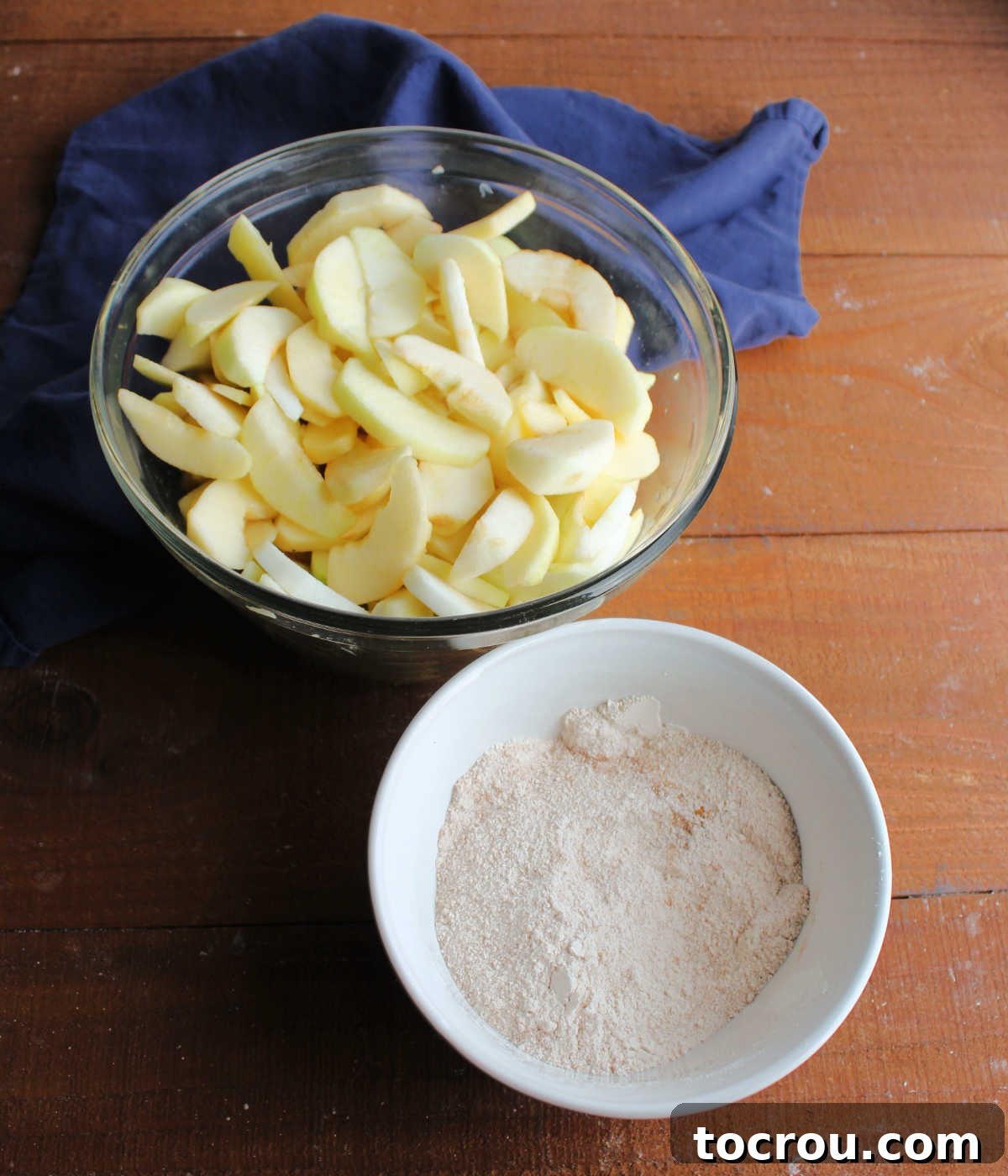 Bowl of thin apple slices tossed in lemon juice next to small bowl of sugar and flour mixed together.