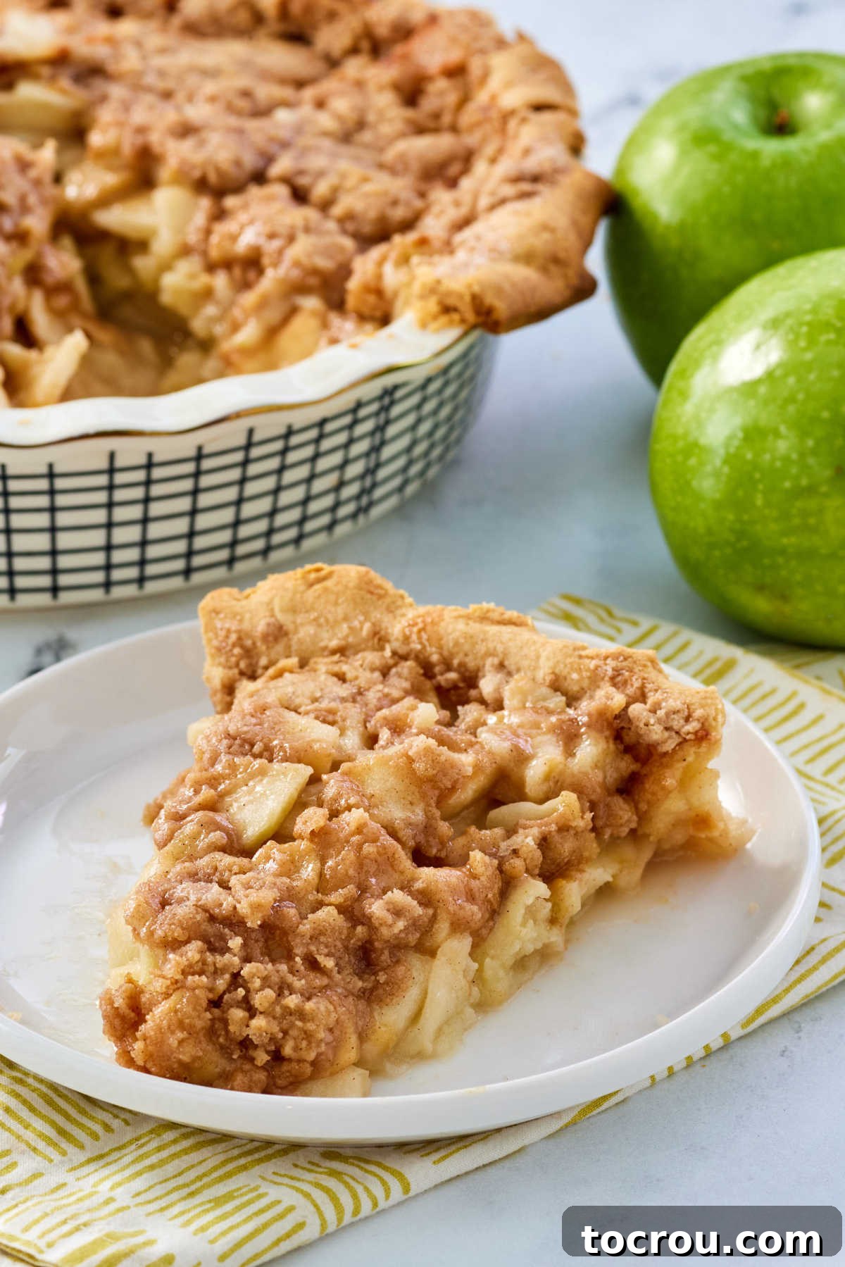Slice of old-fashioned apple pie with crumb topping on plate with remaining pie in the background. 