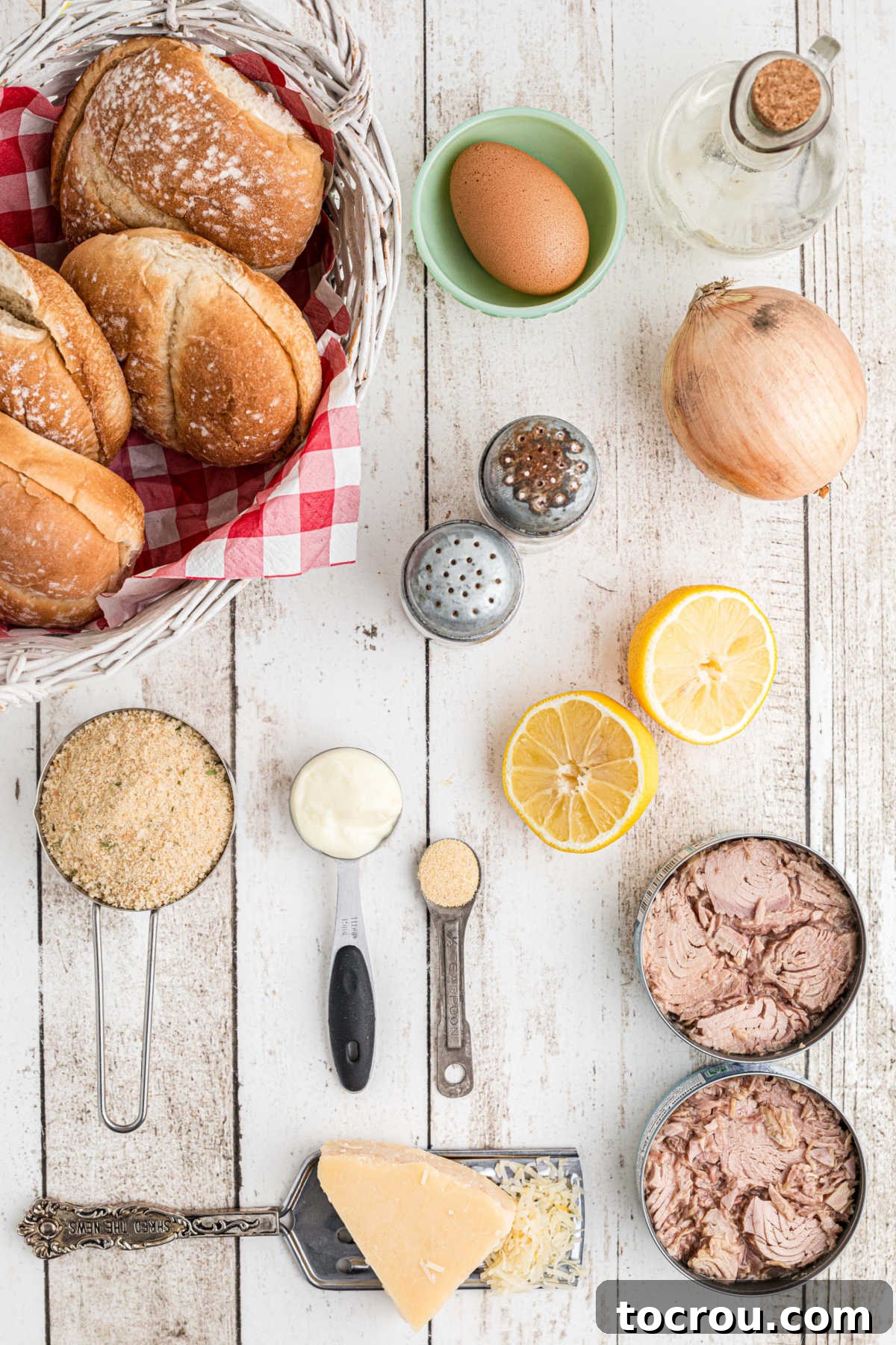 Ingredient shot including cans of tuna, Parmesan cheese, onion, breadcrumbs, lemon, egg, mayonnaise, garlic powder, salt, pepper, and buns.