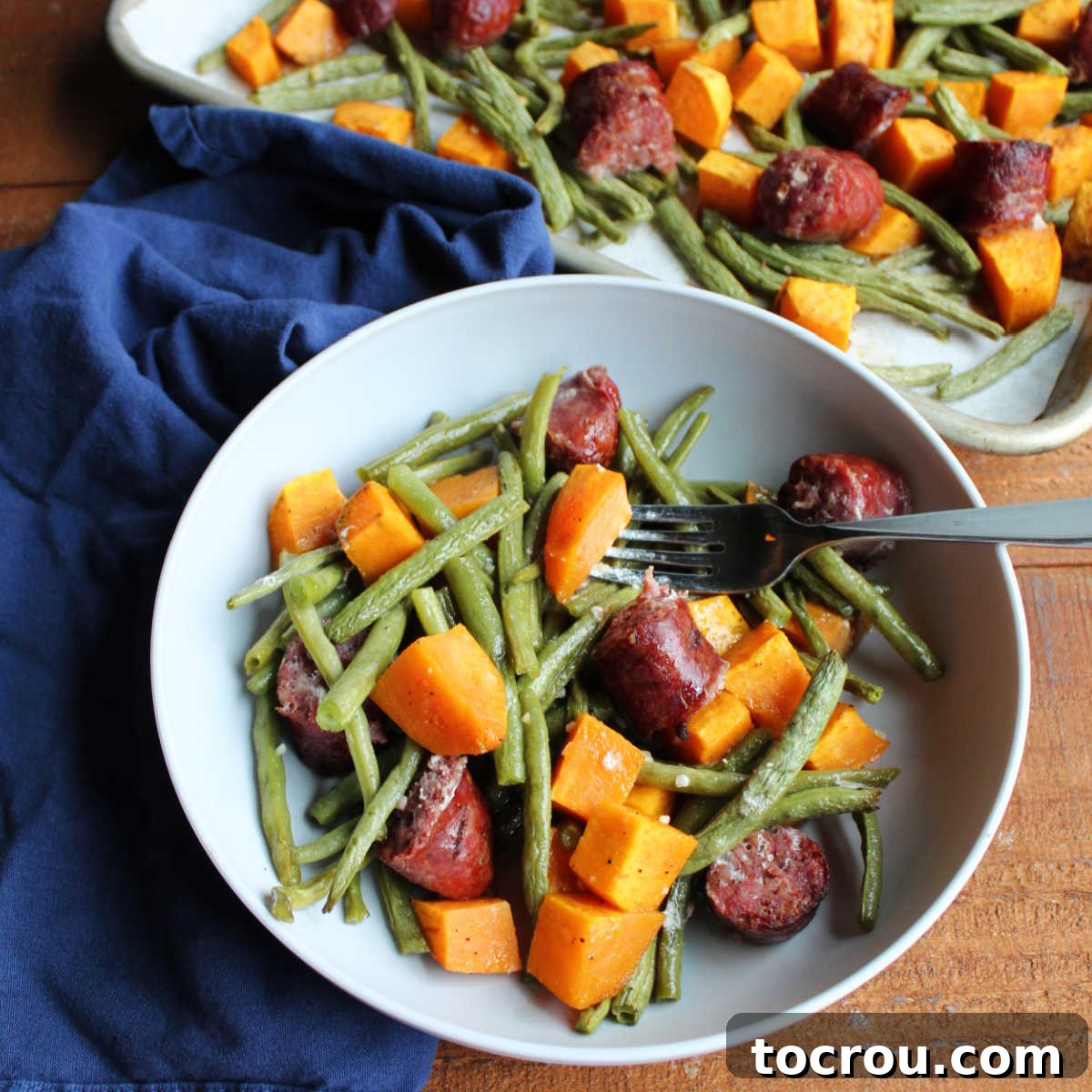 Bowl of roasted green beans, sweet potatoes, and smoked sausage with fork ready to eat and sheet pan with remaining food in background.