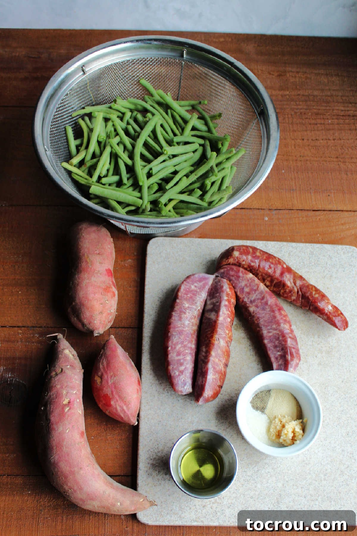 Ingredients including smoked sausage, fresh sweet potatoes and green beans, oil, garlic, salt, pepper, and onion powder ready to be made into sheet pan supper.