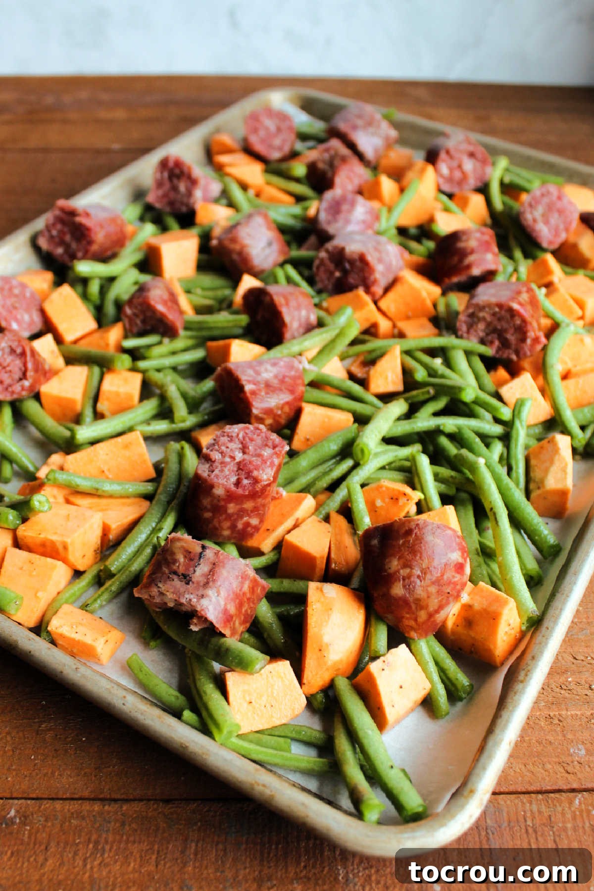 Chunks of sausage placed over green beans and sweet potatoes on sheet pan, ready to go in the oven.