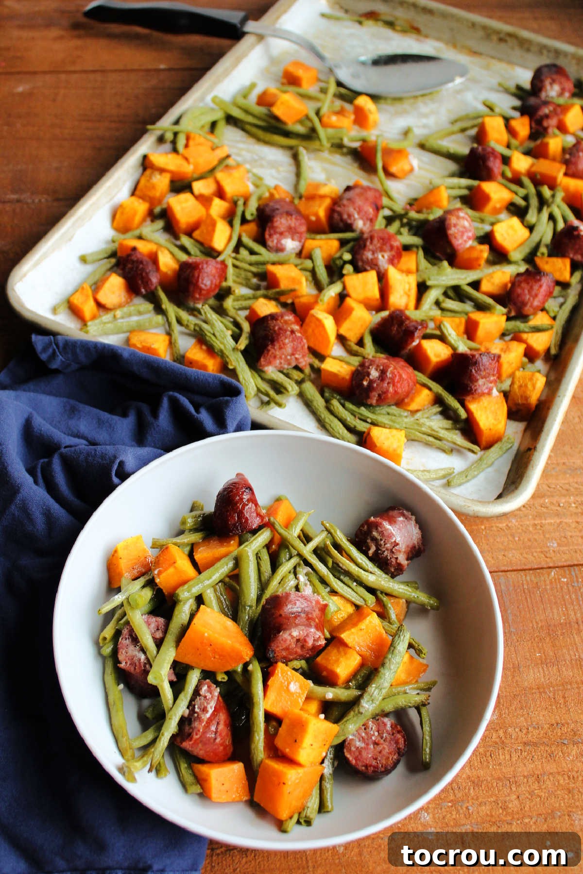 Bowl of roasted sweet potatoes, green beans and sausage ready to eat with sheet pan of remaining food nearby.