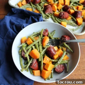 Bowl of roasted green beans, sweet potatoes, and smoked sausage with fork ready to eat and sheet pan with remaining food in background.
