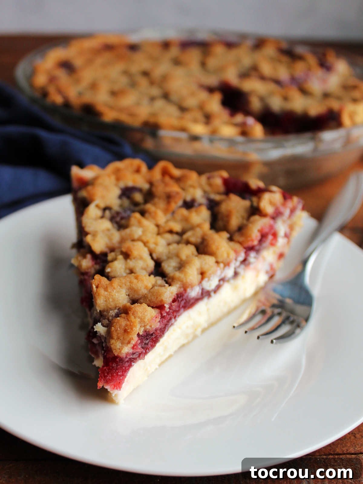Buttery Crumb Cranberry Bliss 11 A beautifully presented slice of cranberry crumble pie, featuring a distinct cream cheese layer, served on a small white plate with a fork, inviting a delicious bite.