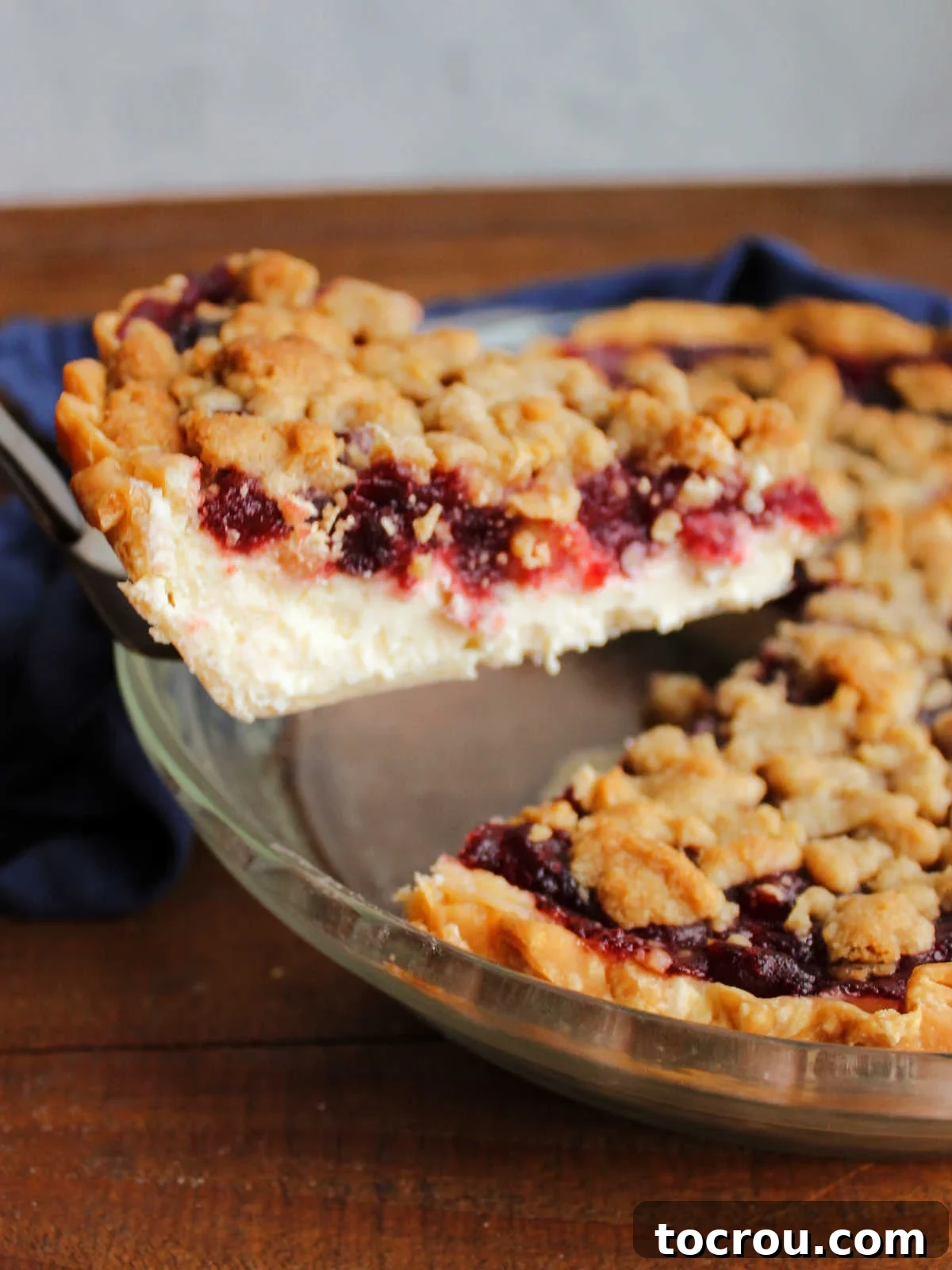 Buttery Crumb Cranberry Bliss 3 A pie server carefully lifting a slice of cranberry crumb pie from the pan, revealing the distinct creamy cheese layer, vibrant cranberry layer, and golden streusel topping.