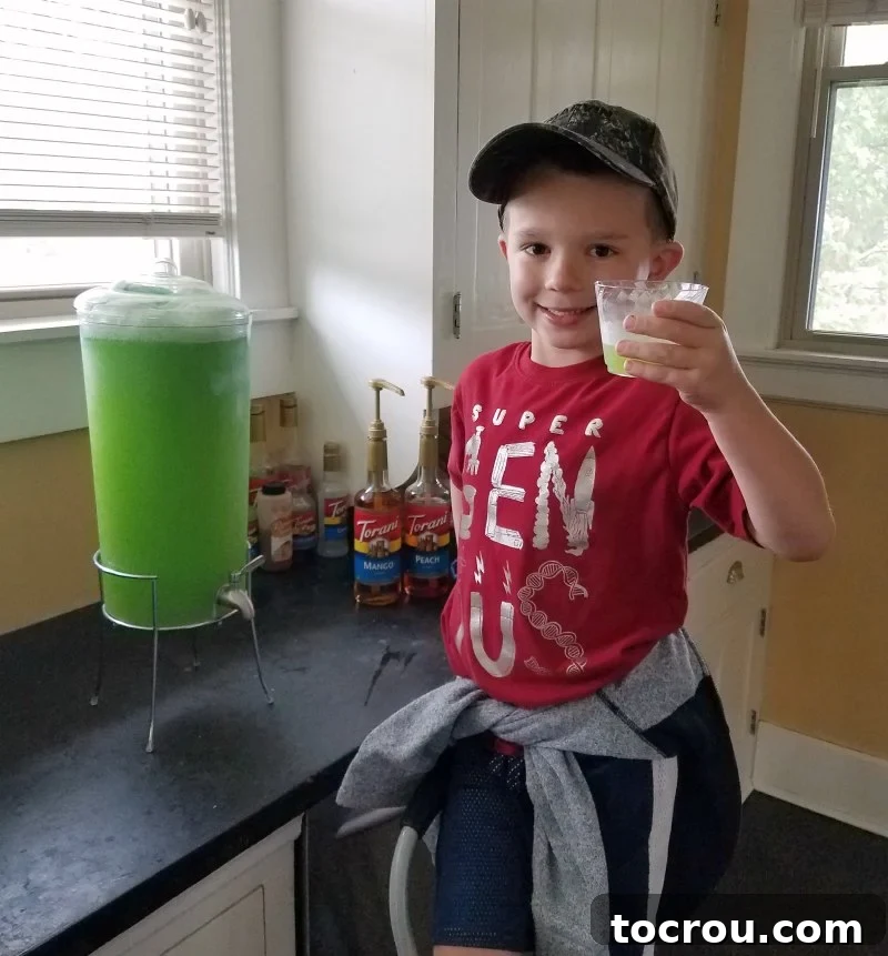Little Dude beaming with pride as he pours the first glass of vibrant green slime punch from the dispenser.