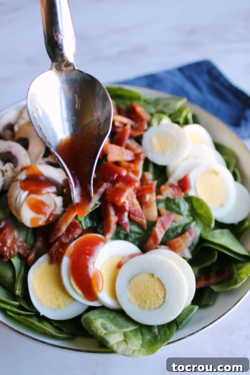 Spinach salad is a favorite for good reason. It is loaded with yummy toppings that work so well with the homemade dressing and spinach base. Serve it as a side or make a meal of it! A drizzle spoon elegantly pouring the vibrant red homemade dressing over a bowl of fresh spinach salad, adorned with perfectly cooked eggs, crispy bacon, and sliced mushrooms.