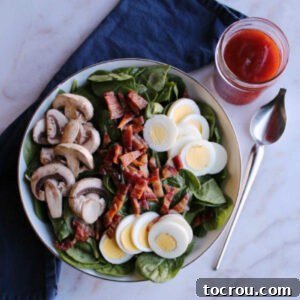 A beautiful bowl of classic spinach salad, featuring crisp spinach, hard-boiled eggs, crumbled bacon, and fresh mushrooms, ready to be dressed and served.