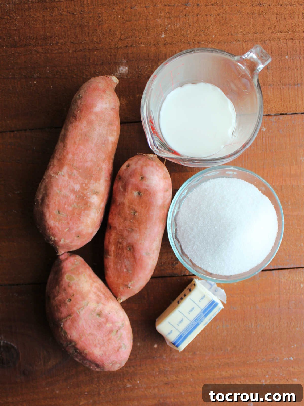 Fresh ingredients laid out: vibrant orange sweet potatoes, a stick of butter, a bowl of granulated sugar, and a glass of milk, ready for cooking.