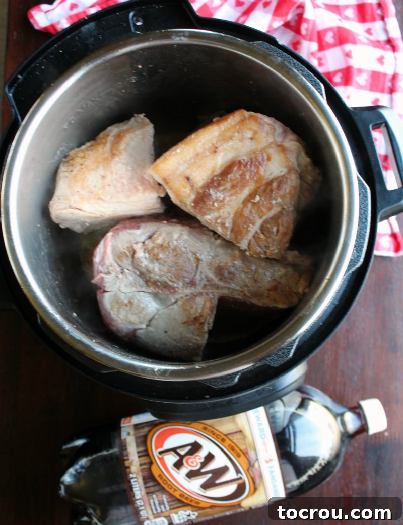 Pork Shoulder Browning in Instant Pot with Root Beer Bottle Pork shoulder pieces browning in an Instant Pot next to a 2-liter bottle of root beer.