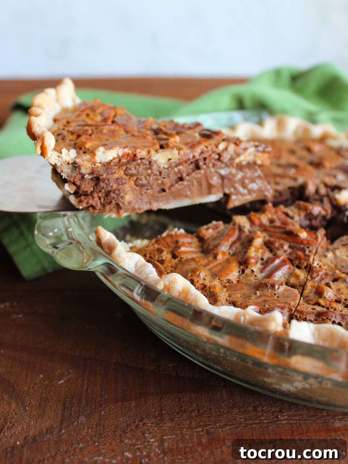 Pie server lifting first piece of chocolate pecan pie out of the pie dish, showing rich chocolate filling and nuts on top.