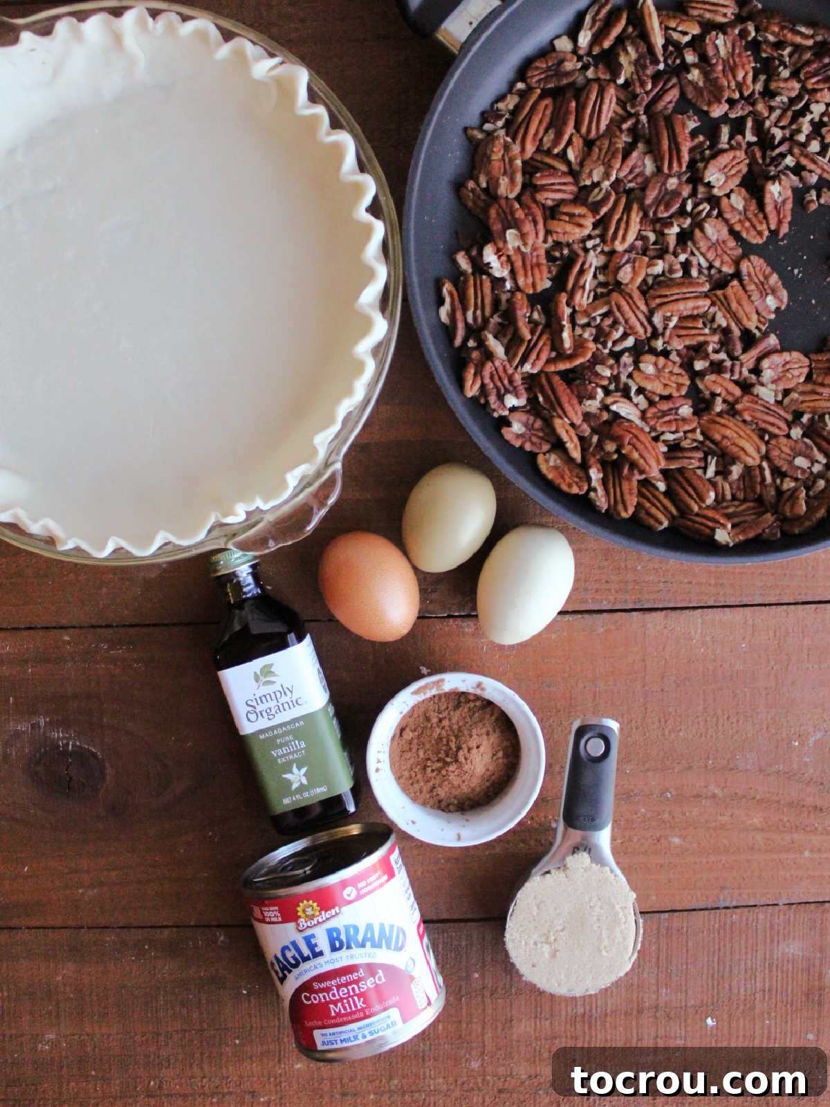 Ingredients including toasted pecans, pie crust, sweetened condensed milk, vanilla, cocoa powder, brown sugar and eggs, ready to be made into chocolate pecan pie.
