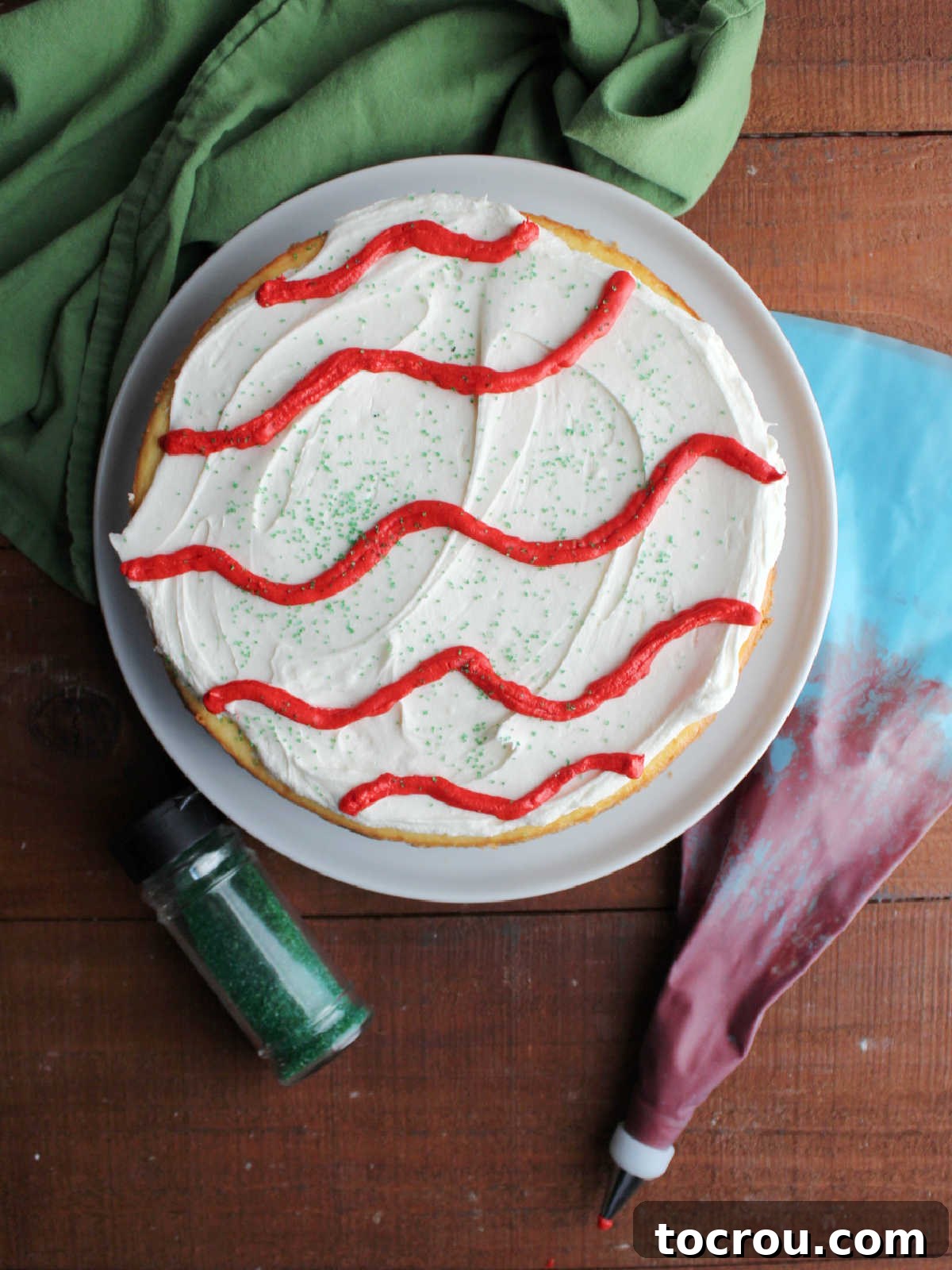 Decorated cheesecake next to piping bag with red frosting and bottle of green sanding sugar.