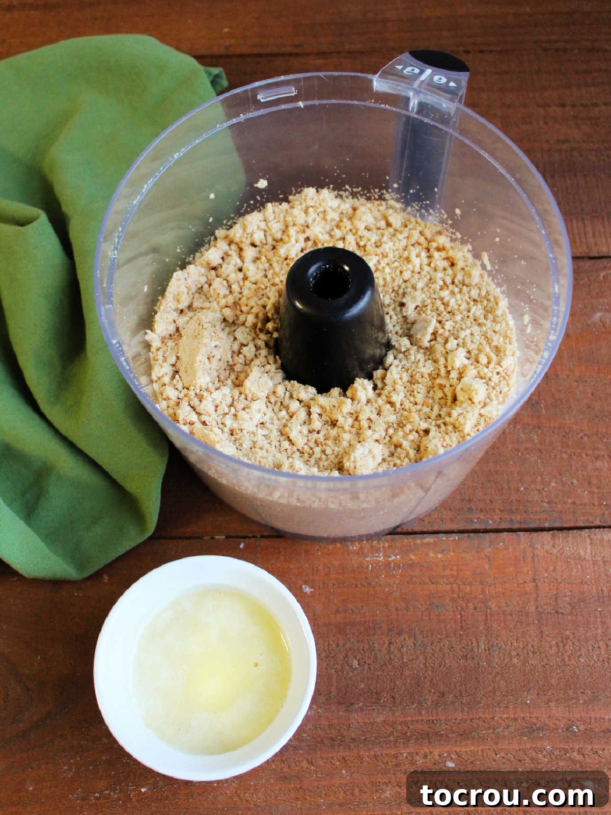 Food processor with Golden Oreo crumbs next to ramekin of melted butter ready to be made into cheesecake crust.