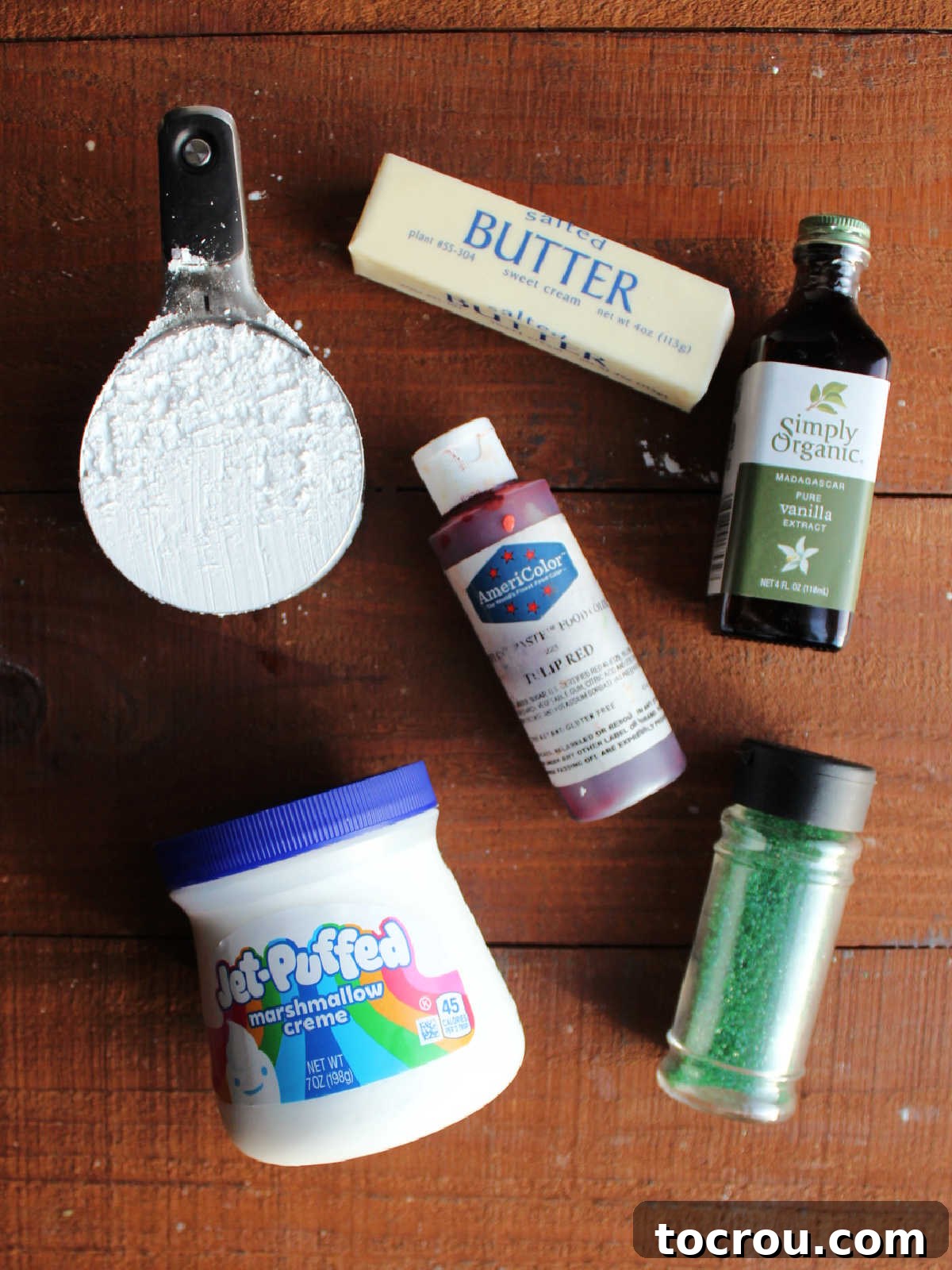 Ingredients including marshmallow fluff, powdered sugar, butter, vanilla, red food coloring and green sanding sugar ready to make frosting and decorate cheesecake.