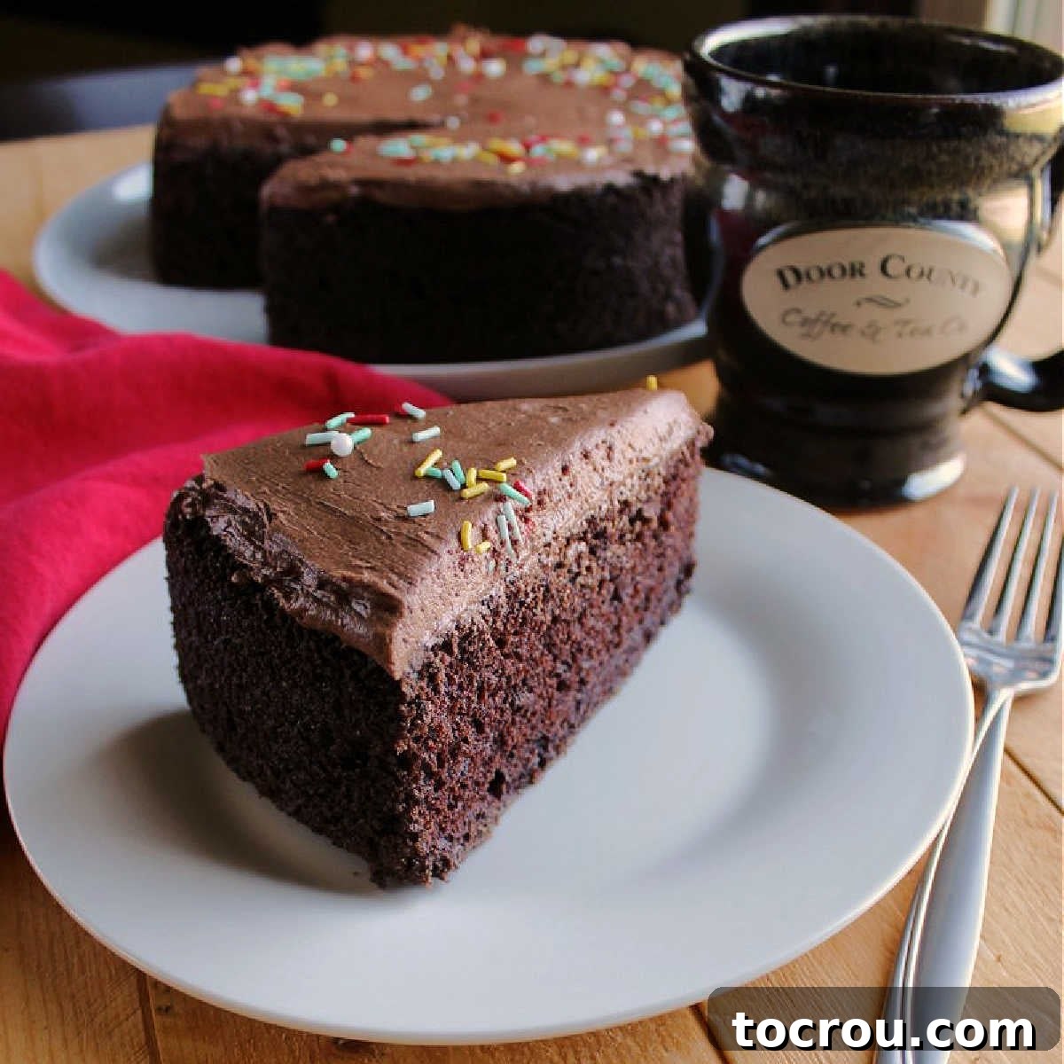 Rich and moist chocolate mayonnaise cake with simple chocolate frosting in a smaller size. It's a perfect dessert for a small party or just because! A perfectly sliced piece of chocolate mayonnaise cake on a white plate, with the remaining cake and a coffee cup blurred in the background, highlighting its moist texture and delicious frosting.