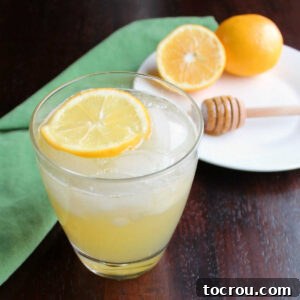 Glass of honey lemonade on ice with a floating lemon slice, accompanied by a honey drizzler and additional fresh lemons in the background.