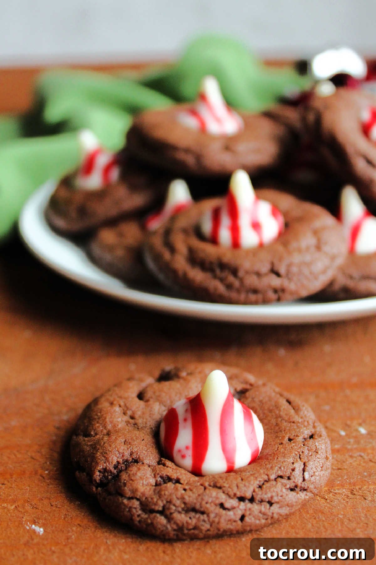 Peppermint Kissed Chocolate Blooms 3 A single chocolate peppermint blossom cookie, showing the soft chocolate base topped with a red and white candy cane Kiss, with more cookies blurred in the background.