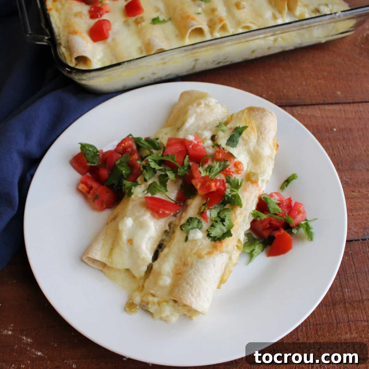 Plate with two breakfast enchiladas baked in creamy cheese sauce and topped with diced tomato and fresh cilantro, ready to eat.