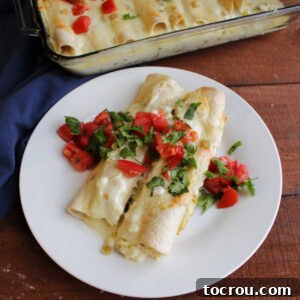Plate with two breakfast enchiladas baked in creamy cheese sauce and topped with diced tomato and fresh cilantro, ready to eat.