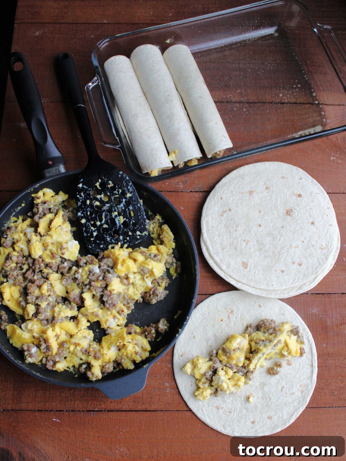Skillet with sausage and egg mixture next to pile of tortillas, one with some sausage and eggs on it and some enchiladas in a baking dish nearby showing how to fill the tortillas and roll them up.