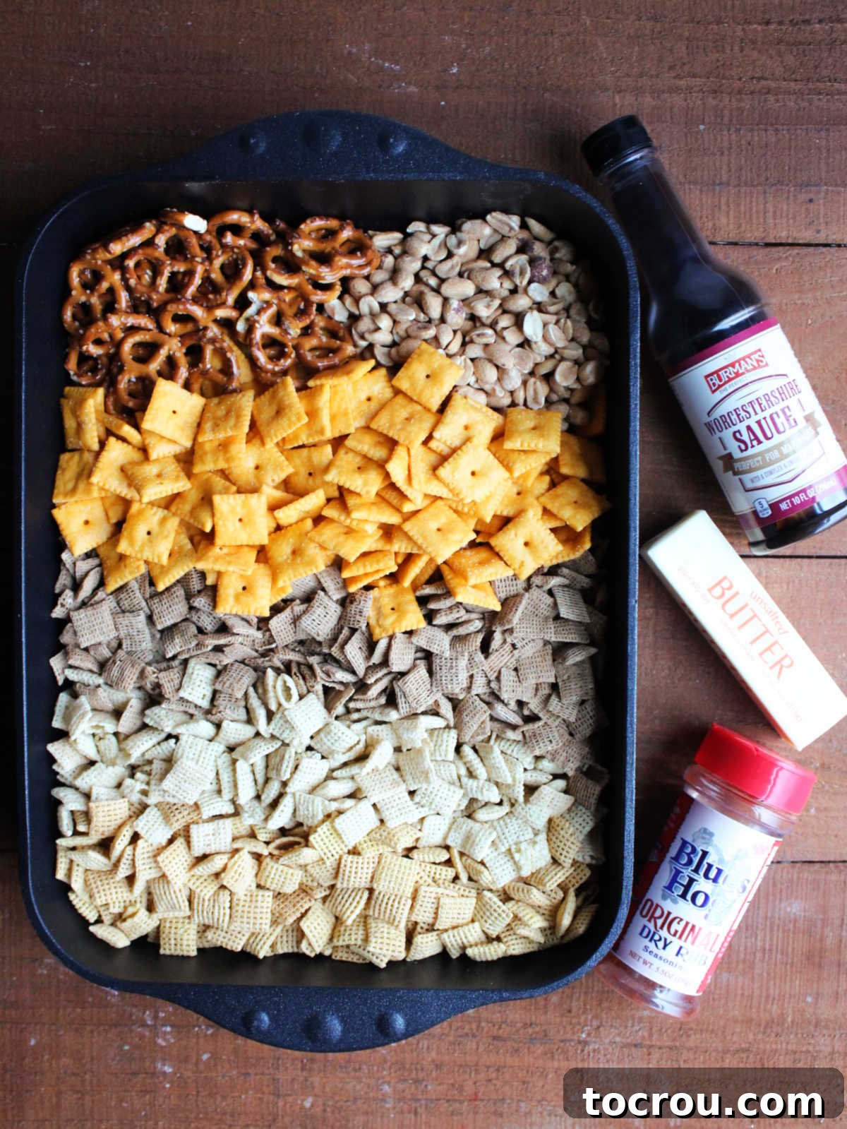 Roasting pan with 3 varieties of Chex cereal, cheese crackers, peanuts, and pretzels inside next to Worcestershire sauce, butter, and Blues Hog BBQ rub, showing ingredients needed to make this snack mix.
