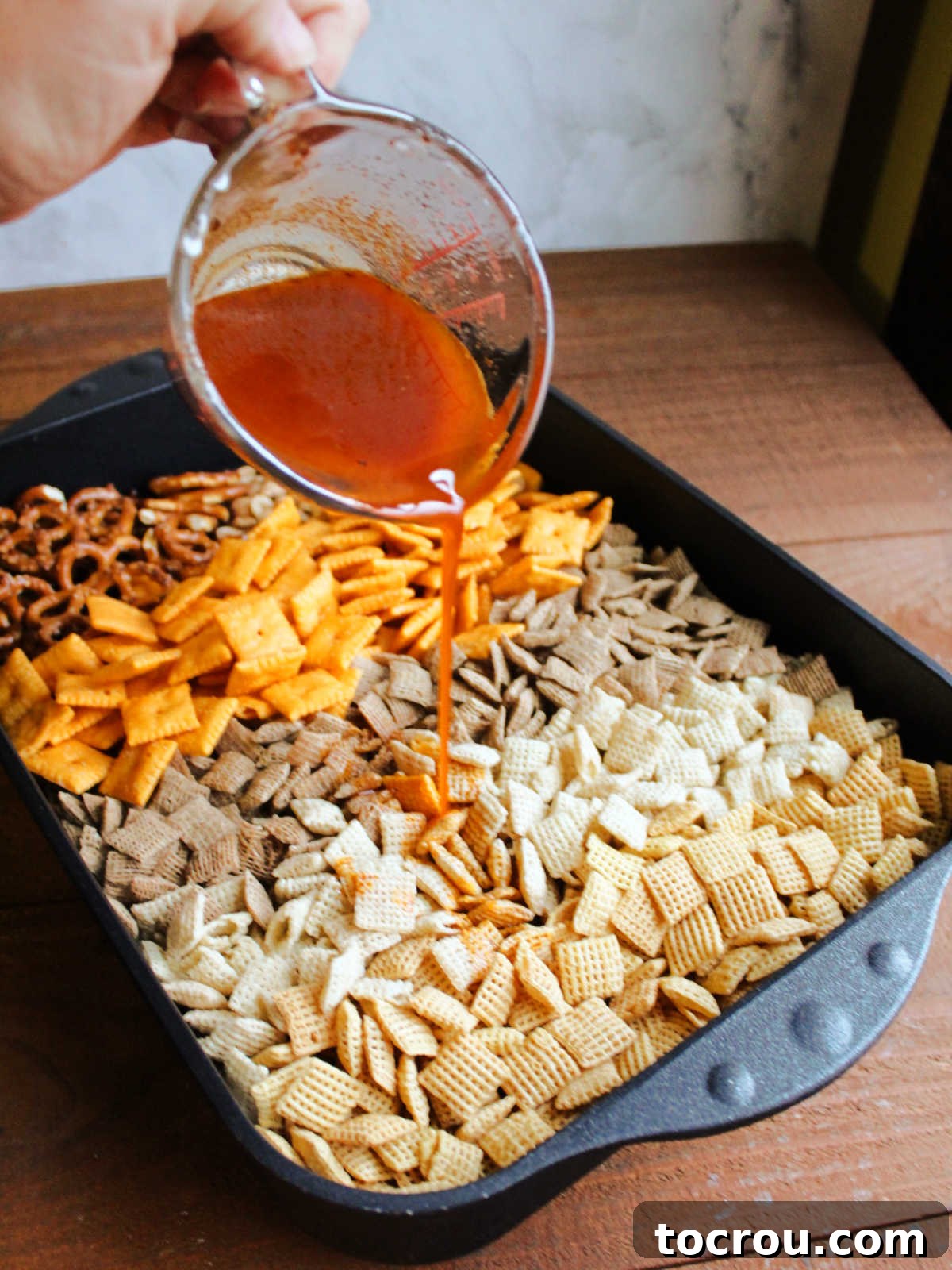 Pouring seasoned butter mixture over a pan of Chex cereal, crackers, pretzels, and peanuts.