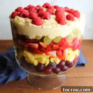 Glass trifle bowl filled with layered fruit salad with red grapes, pineapple, strawberries, green grapes, pudding mixture, and raspberries on top.