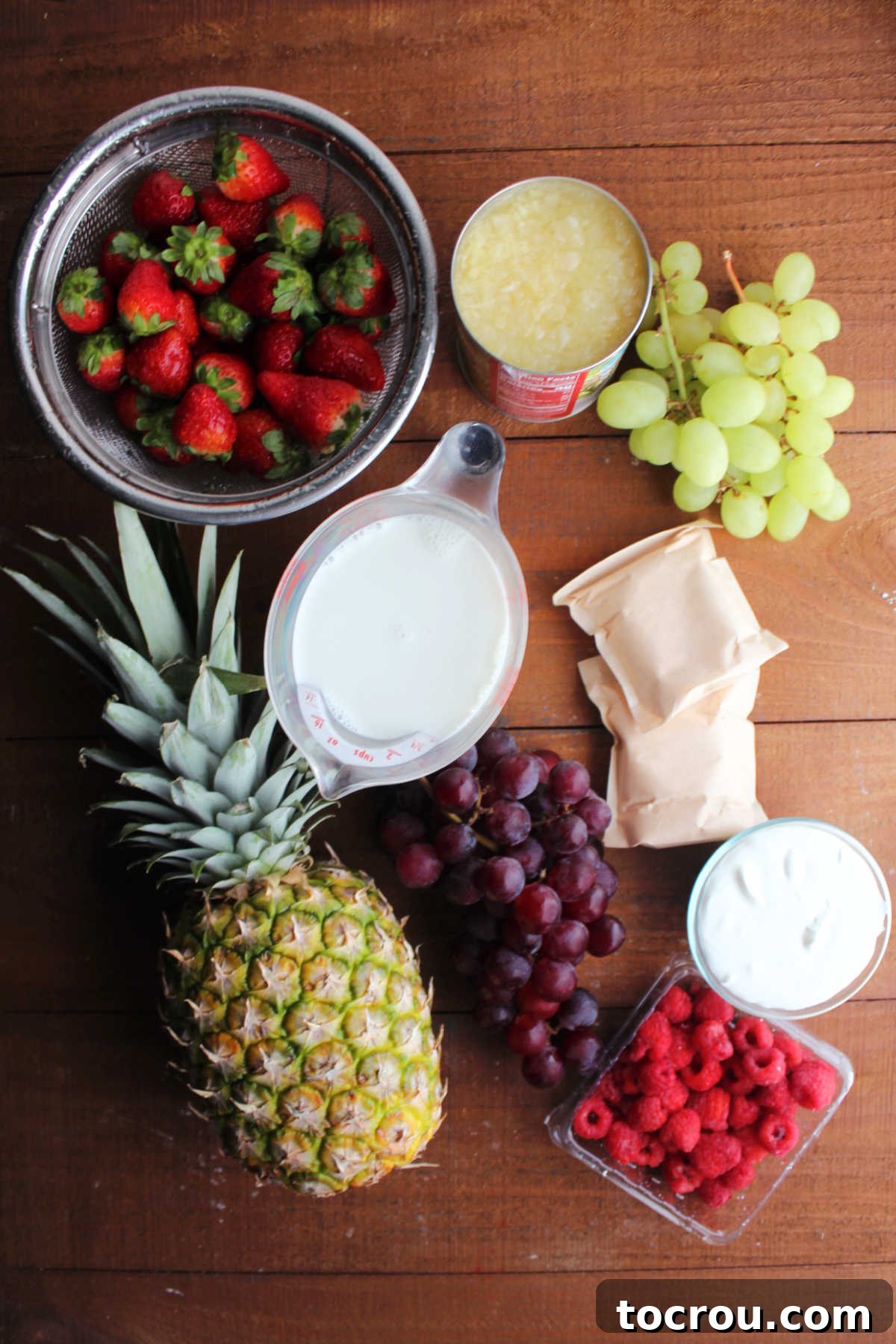 A colorful array of fresh ingredients for fruit salad, including whole pineapples, ripe strawberries, sweet green and red grapes, vibrant raspberries, alongside canned crushed pineapple, instant pudding mix, milk, and sour cream, ready for preparation.