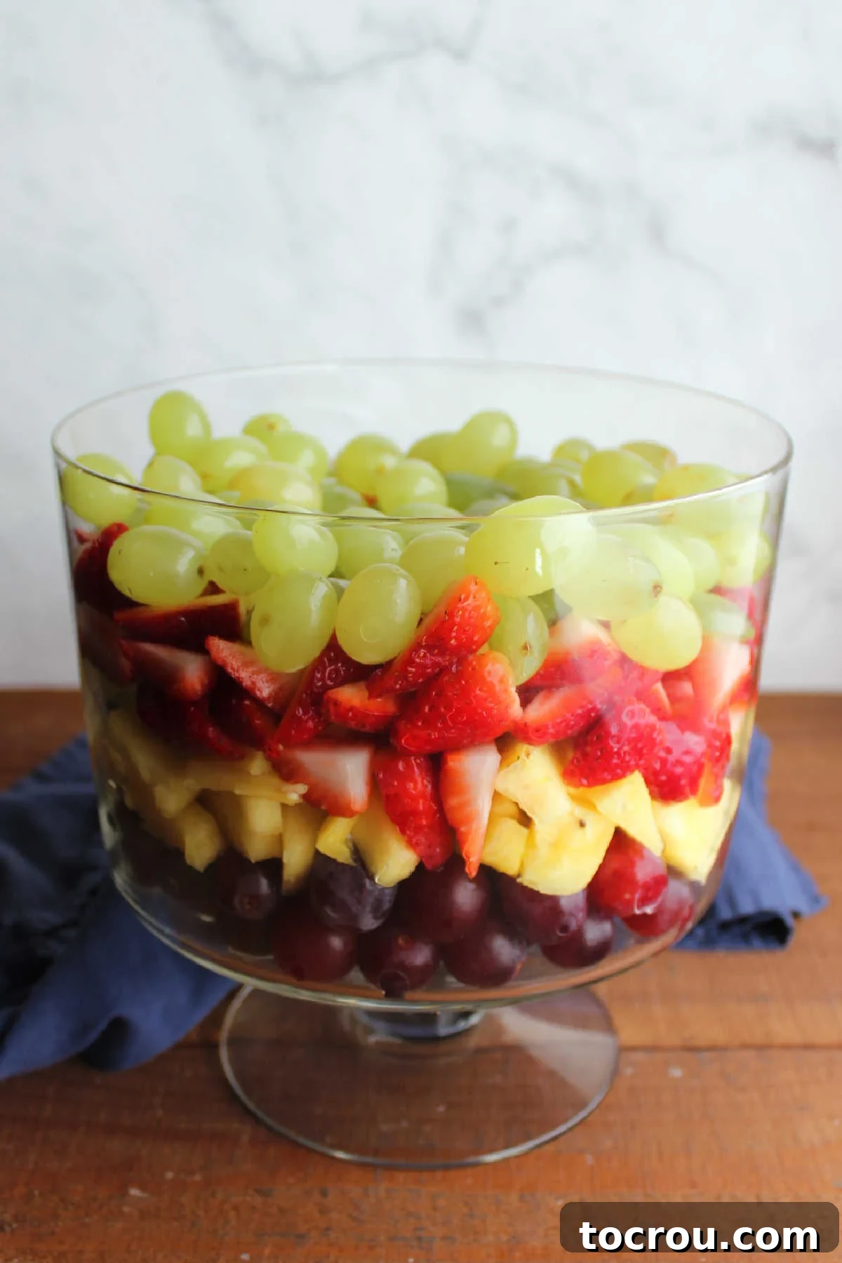 A clear glass trifle bowl showcasing meticulously arranged layers of vibrant red grapes, succulent fresh pineapple chunks, thinly sliced bright strawberries, and plump green grapes, creating a striking visual foundation for the salad.
