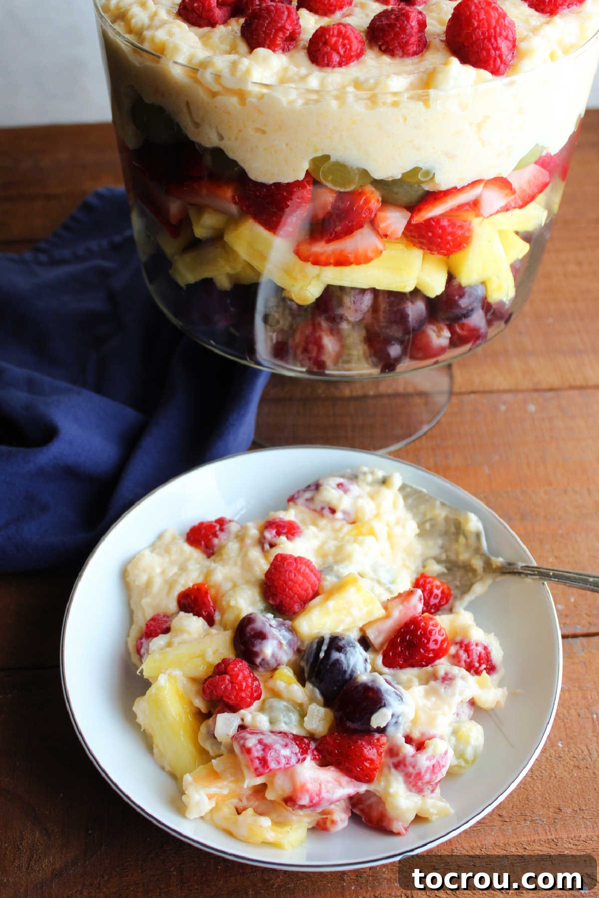 A smaller serving bowl filled with fruit thoroughly coated in the creamy pudding mixture sits beside the main trifle bowl, which still holds the remaining beautifully layered fruit salad.