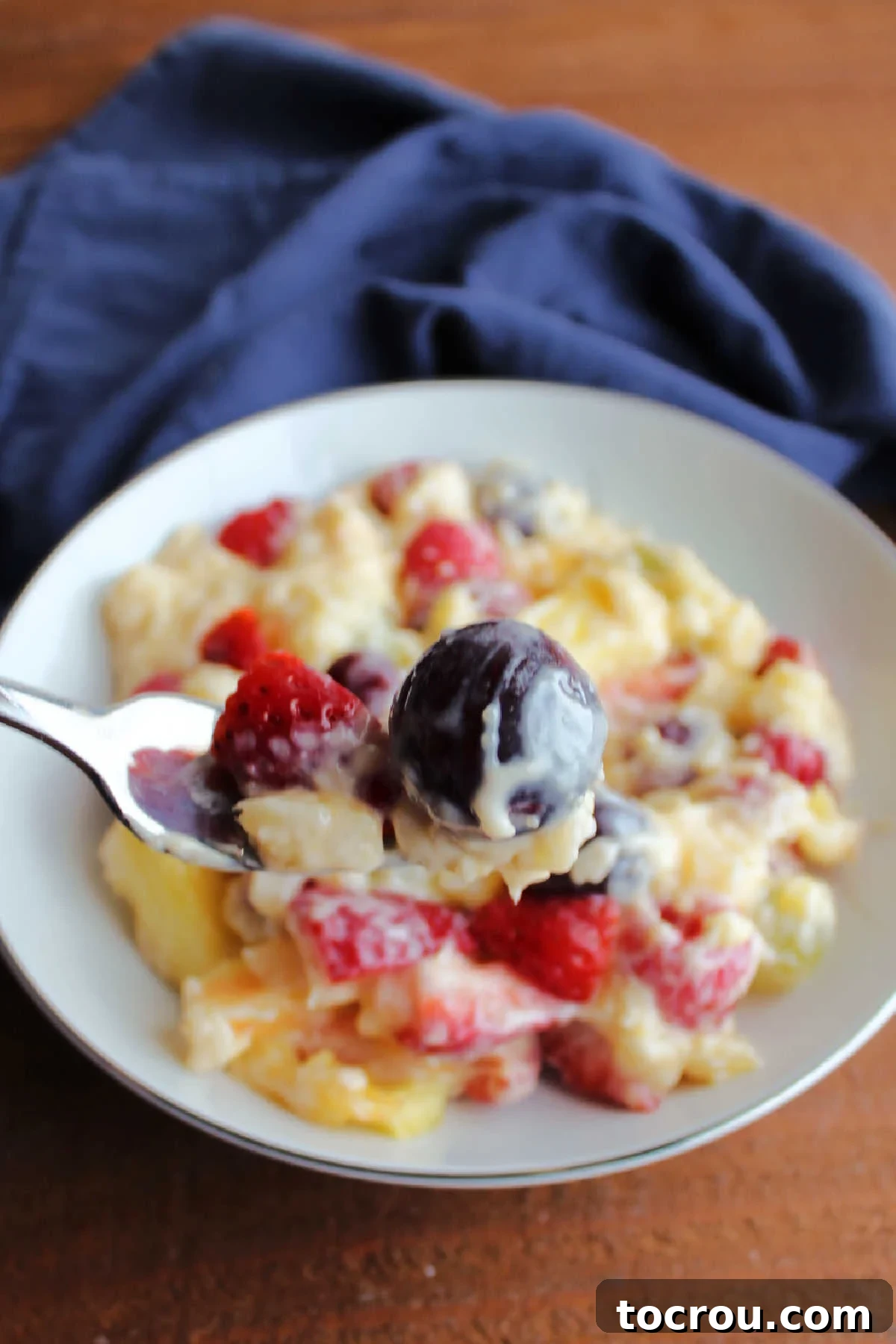 A fork holding a perfect bite of layered fruit salad, featuring a red grape, bright strawberry slices, and a piece of sweet pineapple, all generously coated in the rich, creamy vanilla pudding mixture.
