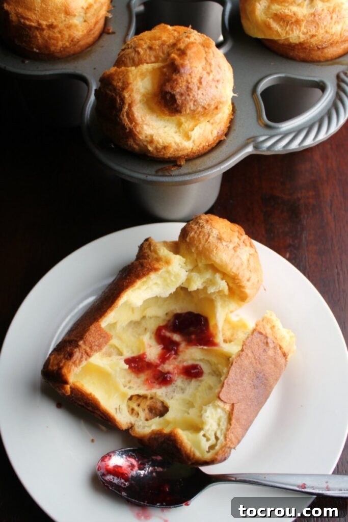 A popover with preserves resting on a plate, with a background shot of a pan filled with large, golden popovers.