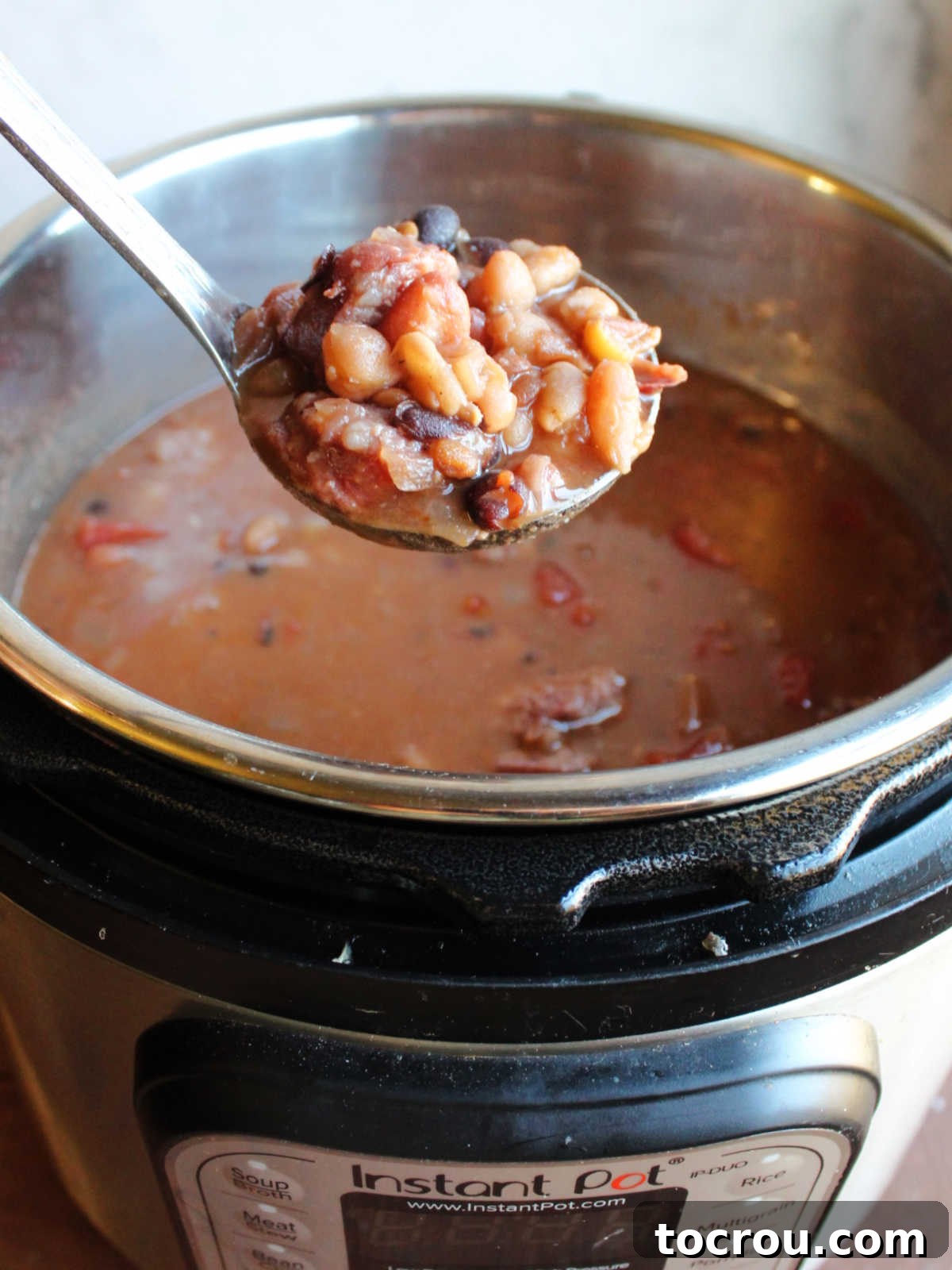 A ladle filled with hearty bean soup and chunks of kielbasa, poised above the Instant Pot containing the rest of the freshly cooked soup, ready to be served.