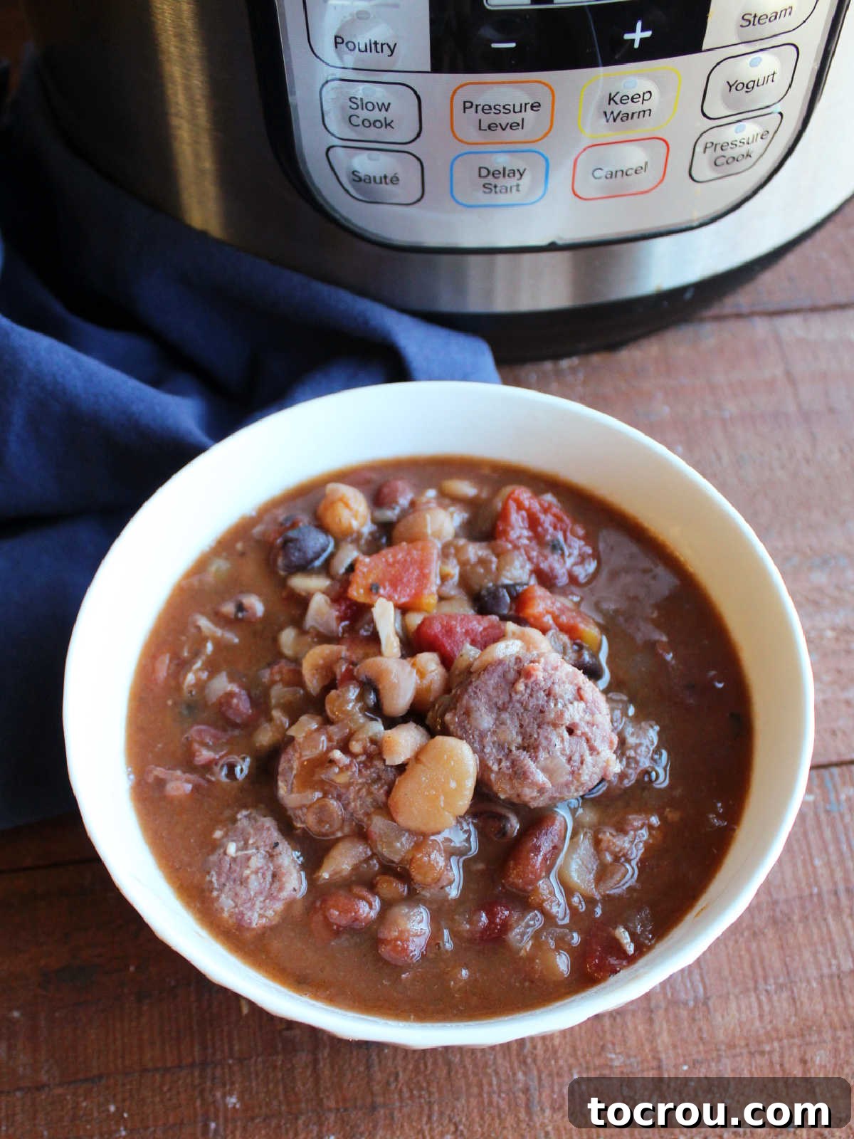 A rustic bowl filled with hearty 16-bean soup and slices of kielbasa, prominently displayed in front of an Instant Pot, suggesting a recently prepared meal.