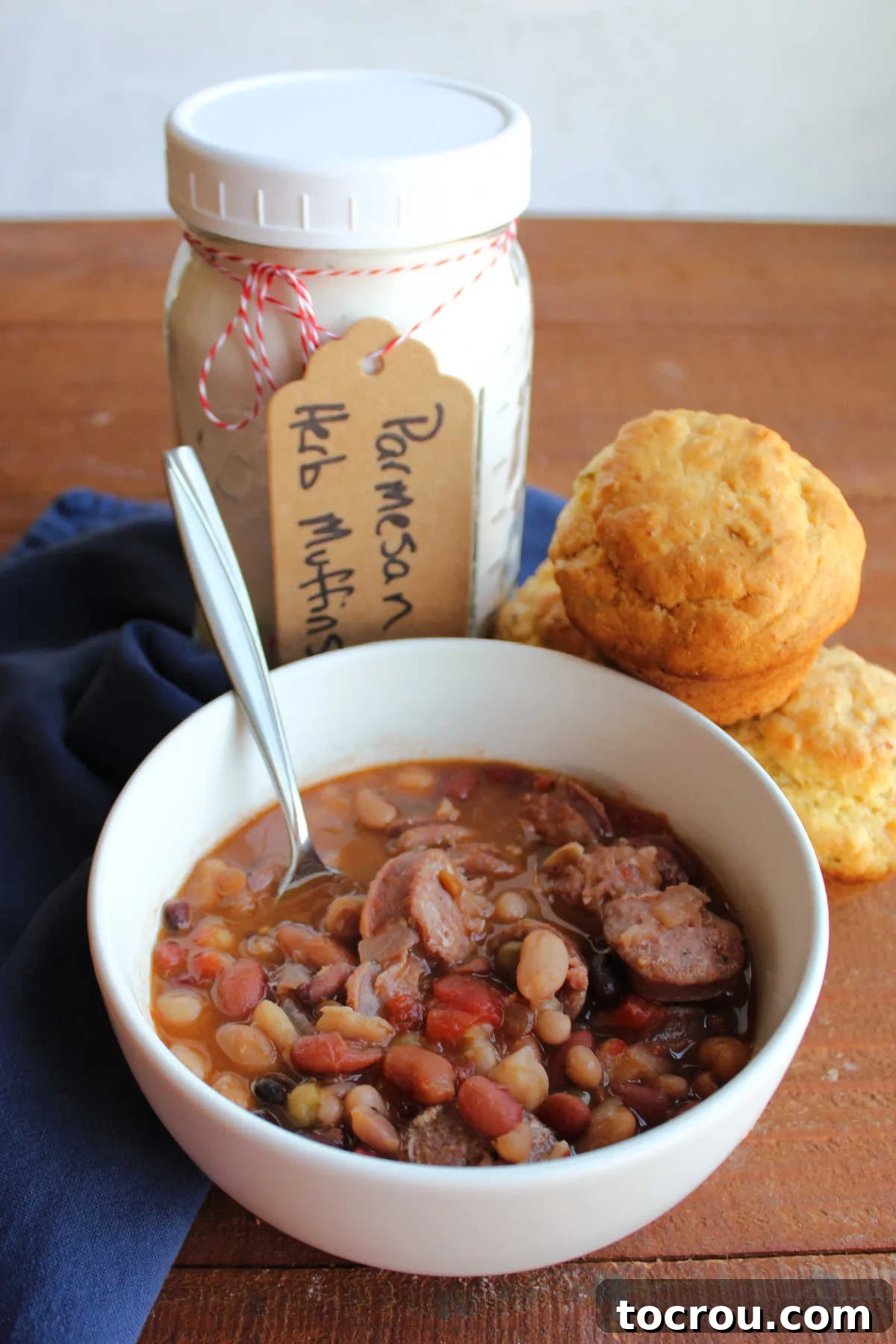 A jar of parmesan muffin mix and a few baked muffins are placed next to a steaming bowl of bean soup with sausage, highlighting complementary serving ideas.