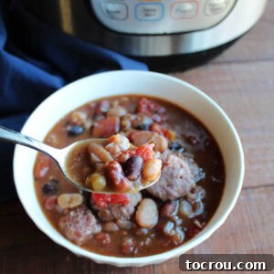 A spoonful of hearty bean soup with tomatoes and kielbasa held over a bowl of soup, next to an Instant Pot.