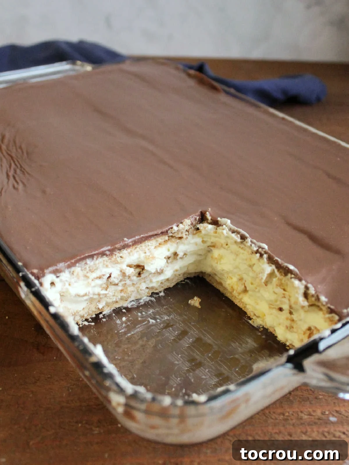Layered Eclair Icebox Cake A panoramic view of a pan of no-bake eclair cake, with a piece removed to showcase the inviting layers of graham crackers, fluffy pudding, and smooth chocolate icing.