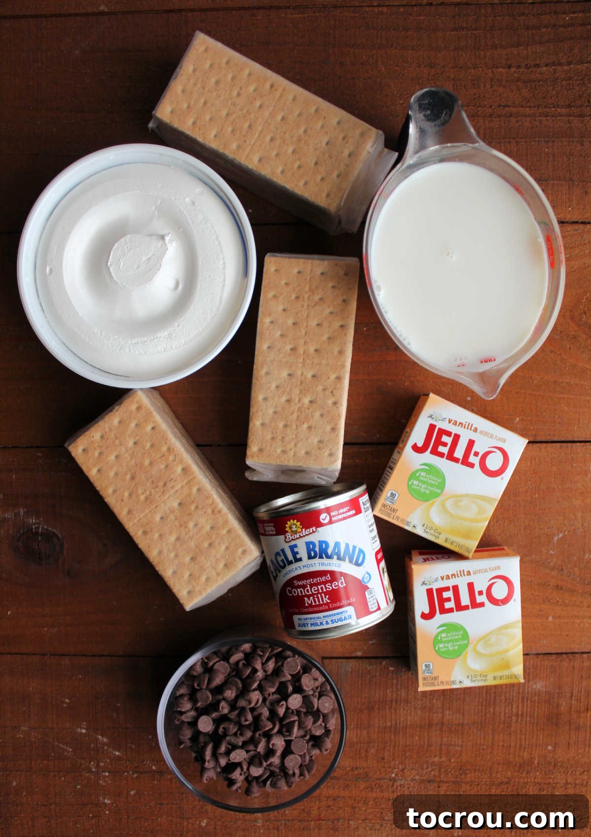 Eclair Cake Ingredients A vibrant display of fresh ingredients for eclair icebox cake, including golden graham crackers, fresh milk, instant vanilla pudding, creamy whipped topping, a can of sweetened condensed milk, and glistening chocolate chips.