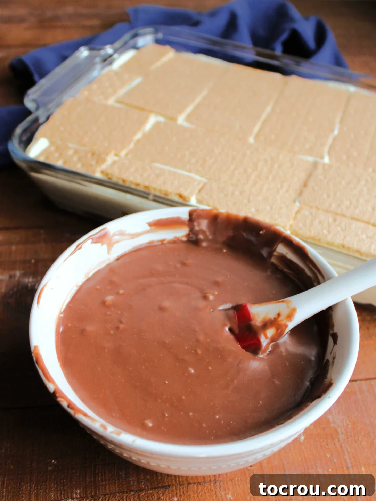 Applying Fudge Icing to Eclair Cake A bowl of freshly melted, warm, and fudgy chocolate icing sitting beside a completed eclair cake in its pan, ready to be spread evenly over the top for a decadent finish.