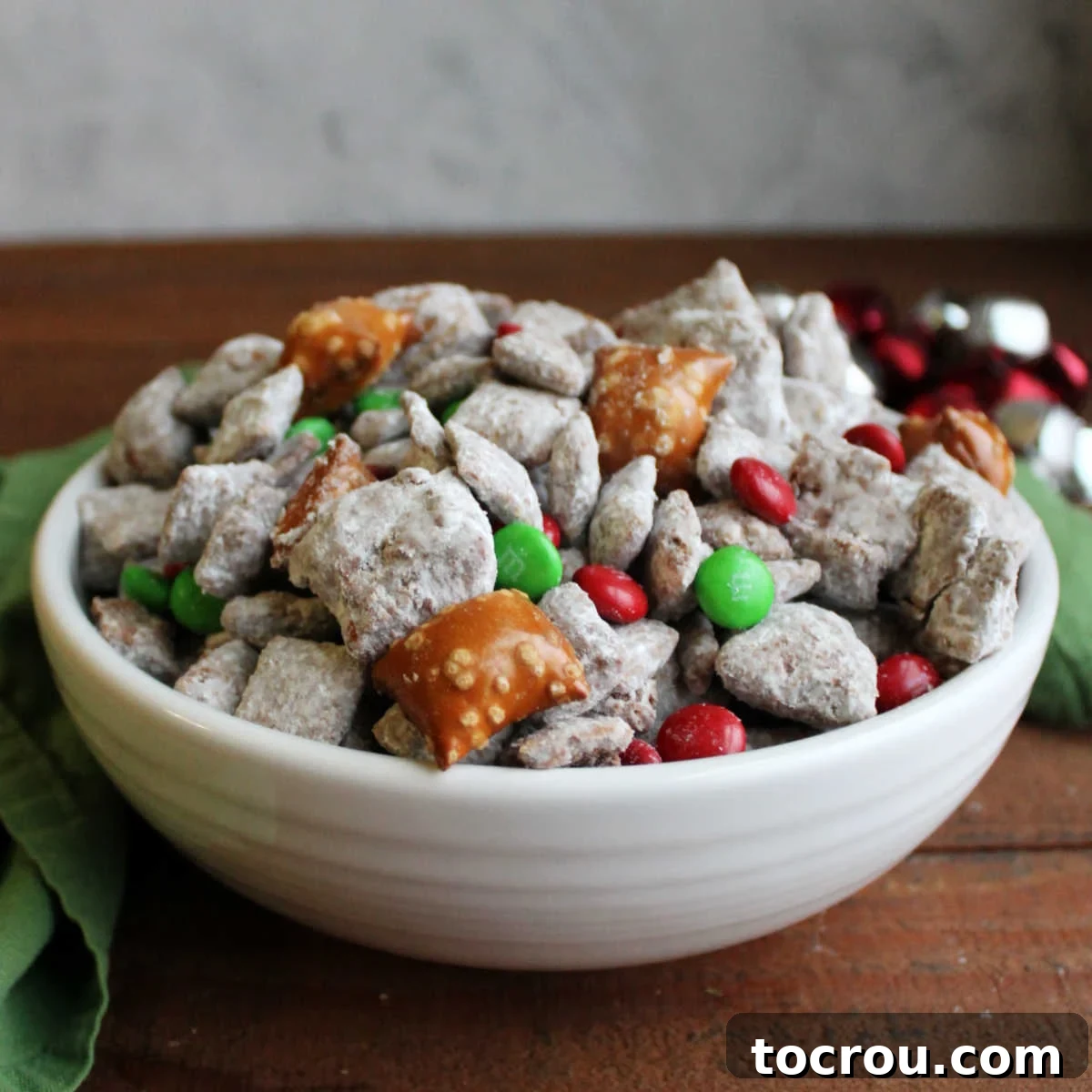 White serving bowl filled with Christmas Chex mix with chocolate and peanut butter muddy buddies, peanut butter filled pretzels, powdered sugar and holiday M&Ms. 