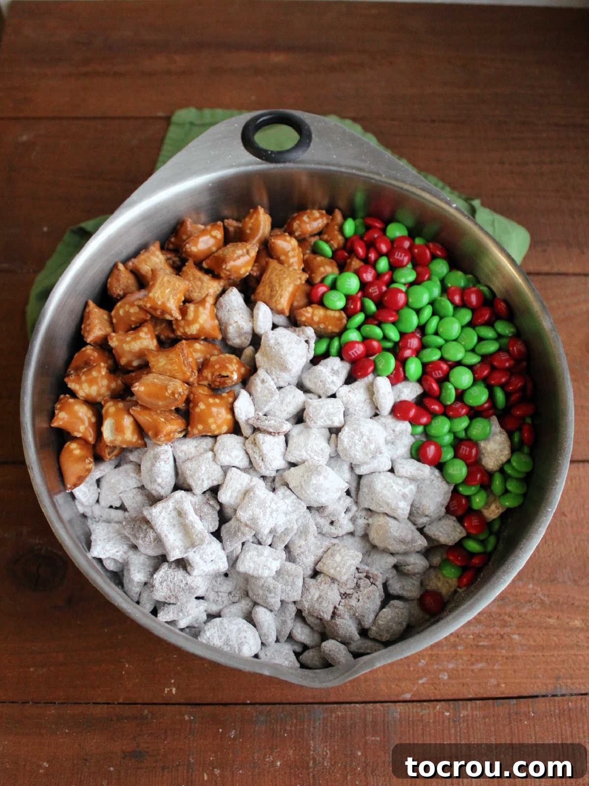 Powdered sugar coated puppy chow in large bowl with holiday M&Ms and more peanut butter filled pretzels. 