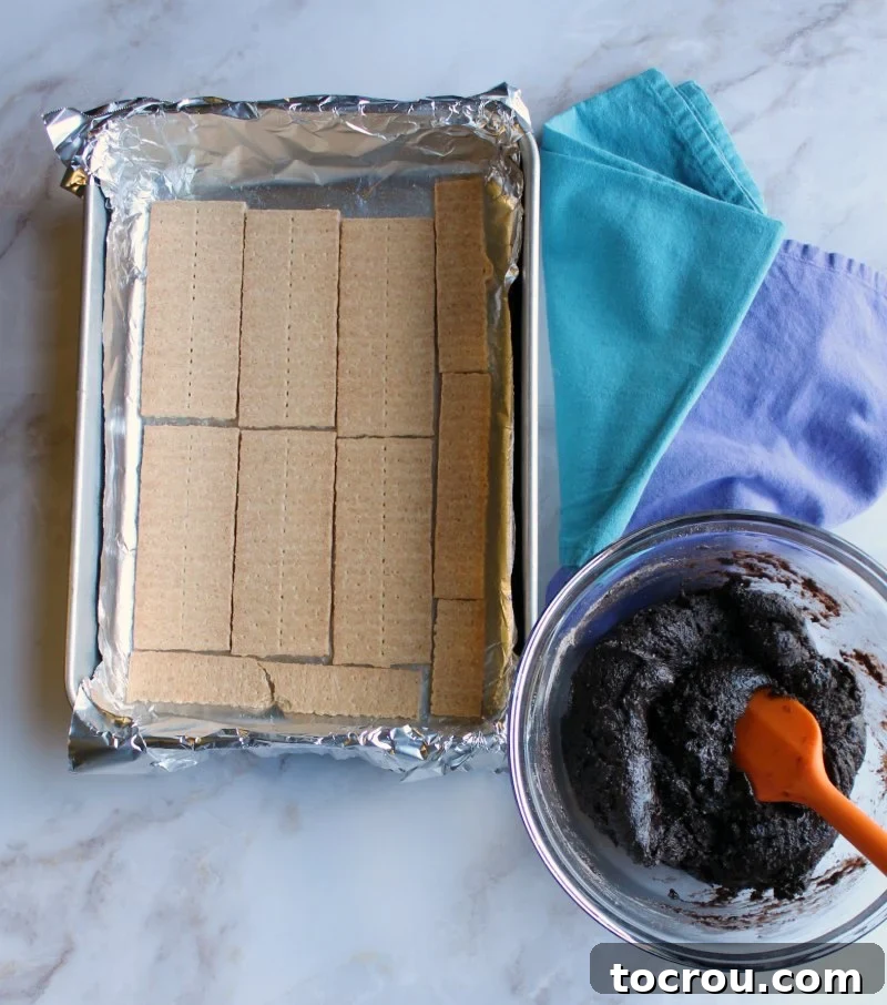 Graham cracker base in a baking pan. Graham crackers carefully placed in a foil-lined baking pan, next to a bowl of chocolate batter.