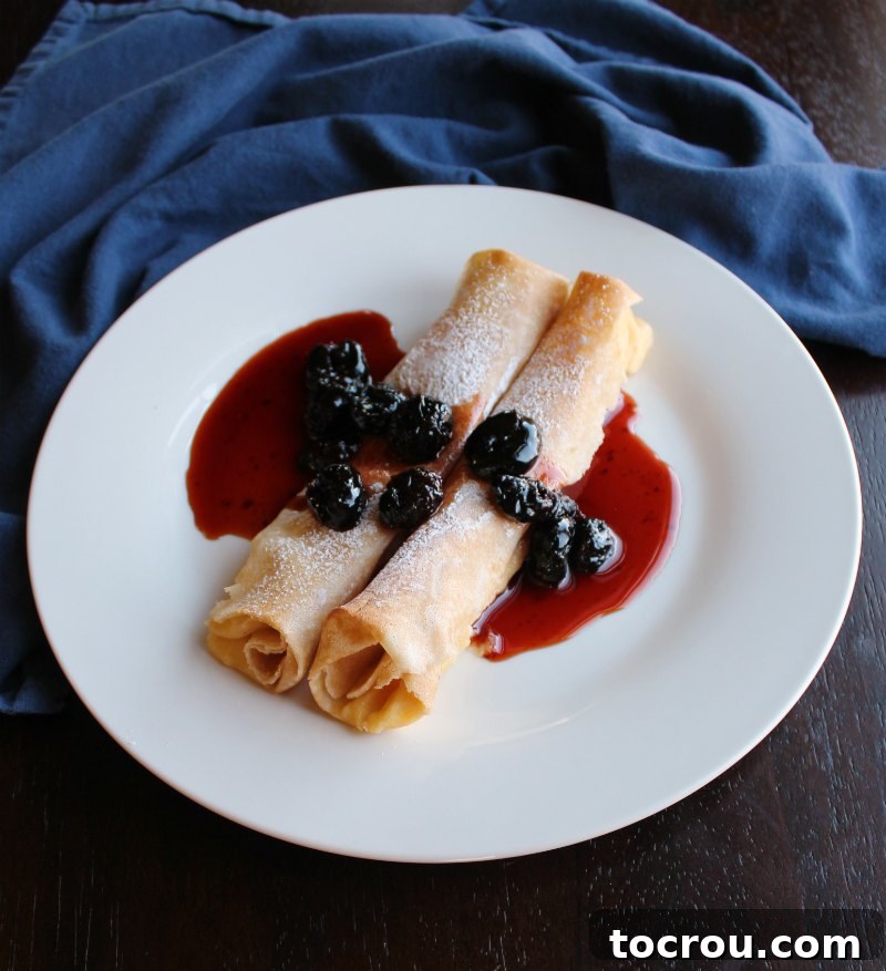 Plate with two rolled crepes filled with lemon ricotta filling and topped with powdered sugar and cherry sauce.