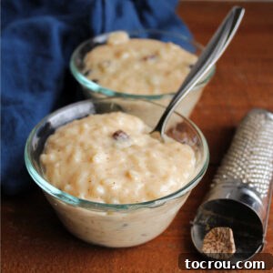Two small glass bowls of creamy condensed milk rice pudding with golden raisins next to a traditional nutmeg grater.
