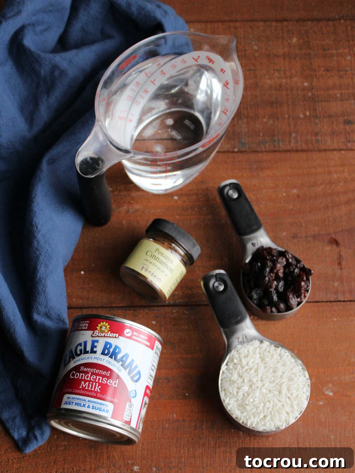 Ingredients including water, a can of sweetened condensed milk, uncooked long-grain rice, a handful of raisins, and ground cinnamon, all ready for making stovetop rice pudding.