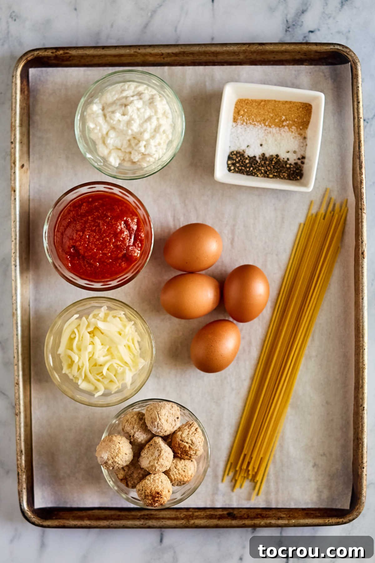 Ingredients including spaghetti noodles, eggs, cottage cheese, marinara sauce, meatballs, mozzarella cheese, and seasonings ready to be made into baked spaghetti and meatballs.