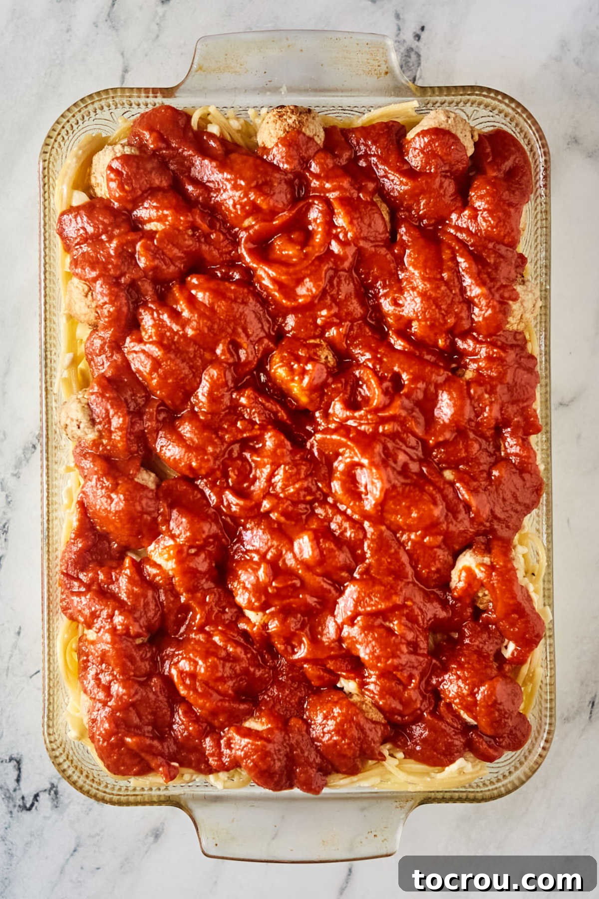 Marinara sauce distributed over the meatballs on top of cheesy spaghetti mixture in casserole dish.