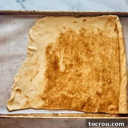 Doug with melted cinnamon sugar butter spread over it, starting to roll it up to form cinnamon rolls.